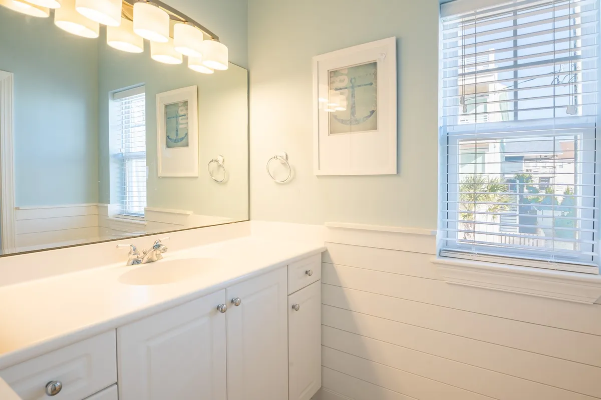 A bright, inviting bathroom with a vanity mirror, light fixtures, a window with blinds, and framed artwork. The scene conveys a serene, well-equipped space for daily routines and relaxation.