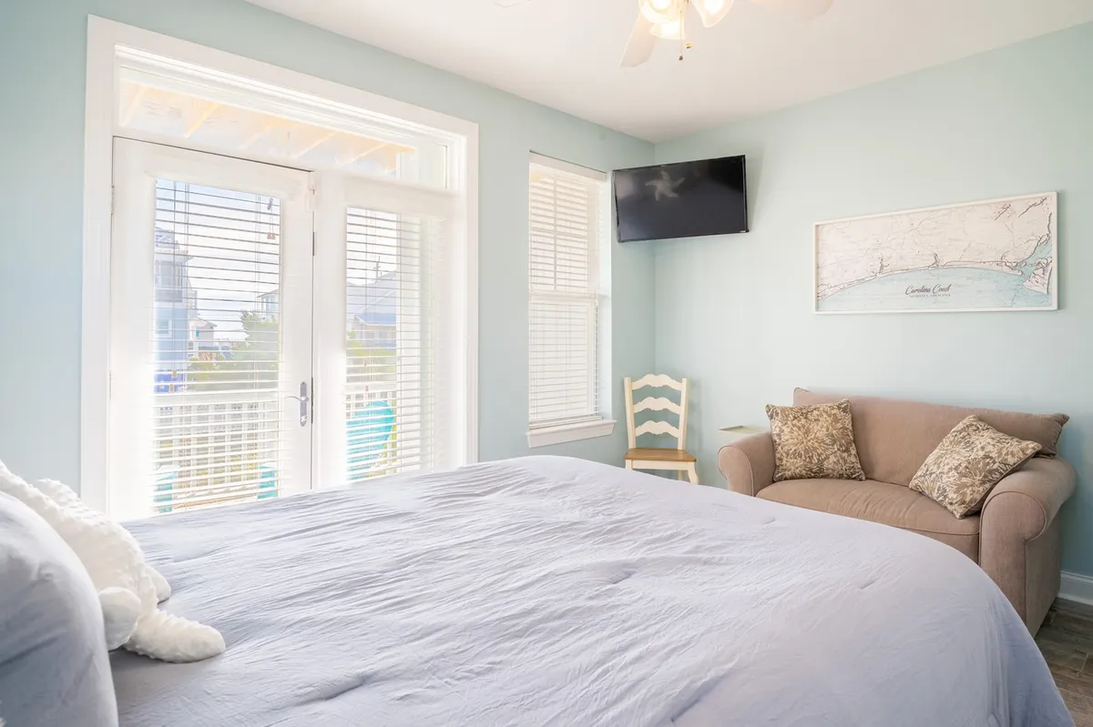 A calm, peaceful bedroom with a comfortable bed, a cozy couch, window views, a TV, and tasteful wall art. The natural light creates an airy feel, ideal for guests looking to unwind, watch a movie, or enjoy a quiet moment with a book.