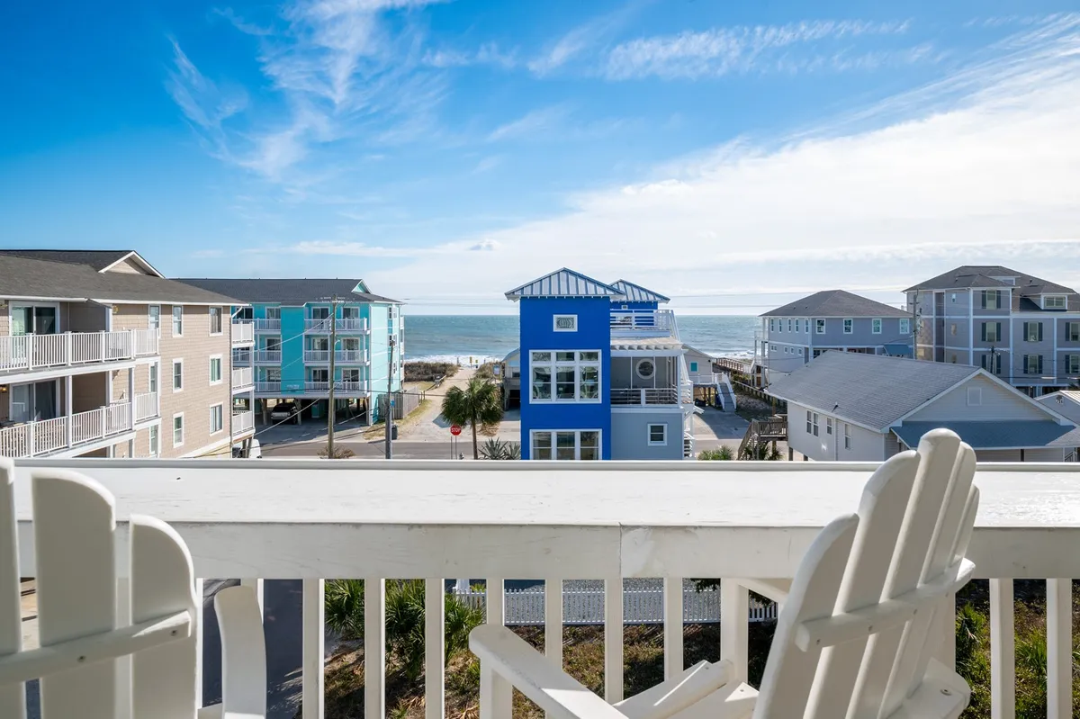 A balcony scene showing two chairs overlooking the ocean, with a blue house in view. The calm setting invites guests to enjoy sunrise coffee or evening relaxation while being immersed in local beach life.