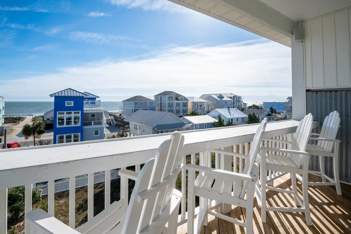 A stunning water-facing view with white rocking chairs on a balcony overlooking the ocean and blue houses in the distance. The scene radiates comfort, peacefulness, and the quintessential coastal retreat atmosphere.