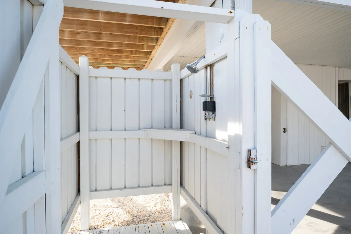 This exterior image features an outdoor shower area framed by a white wooden fence and bathed in sunlight. The space is clean, refreshing, and clearly designed for rinsing off after beach play, with ample room for family members and their pets to use together.