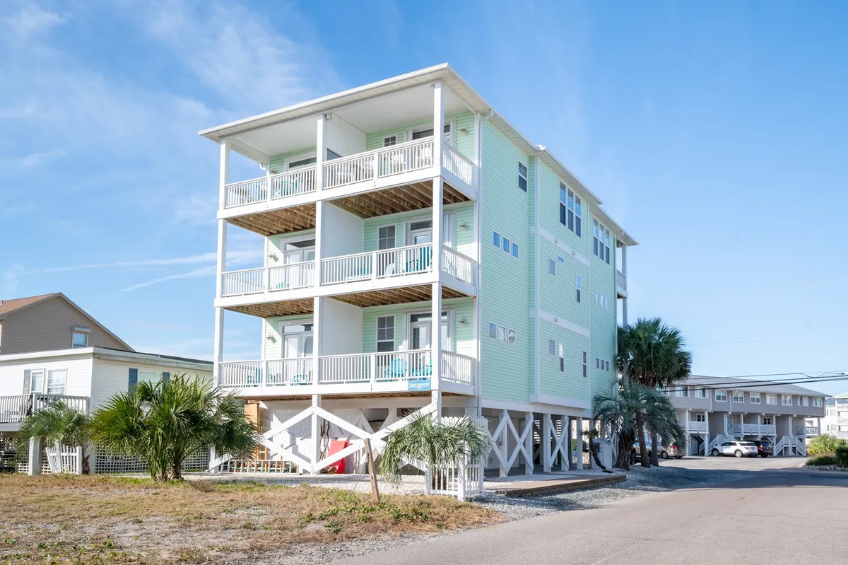 The image showcases a cheerful, three-story beach house with multiple covered balconies, swaying palm trees, and a welcoming driveway. The balconies offer panoramic water views, perfect for soaking in colorful sunsets or enjoying morning coffee. The home’s proximity to Carolina Beach is suggested by the beachy landscaping and easy access.