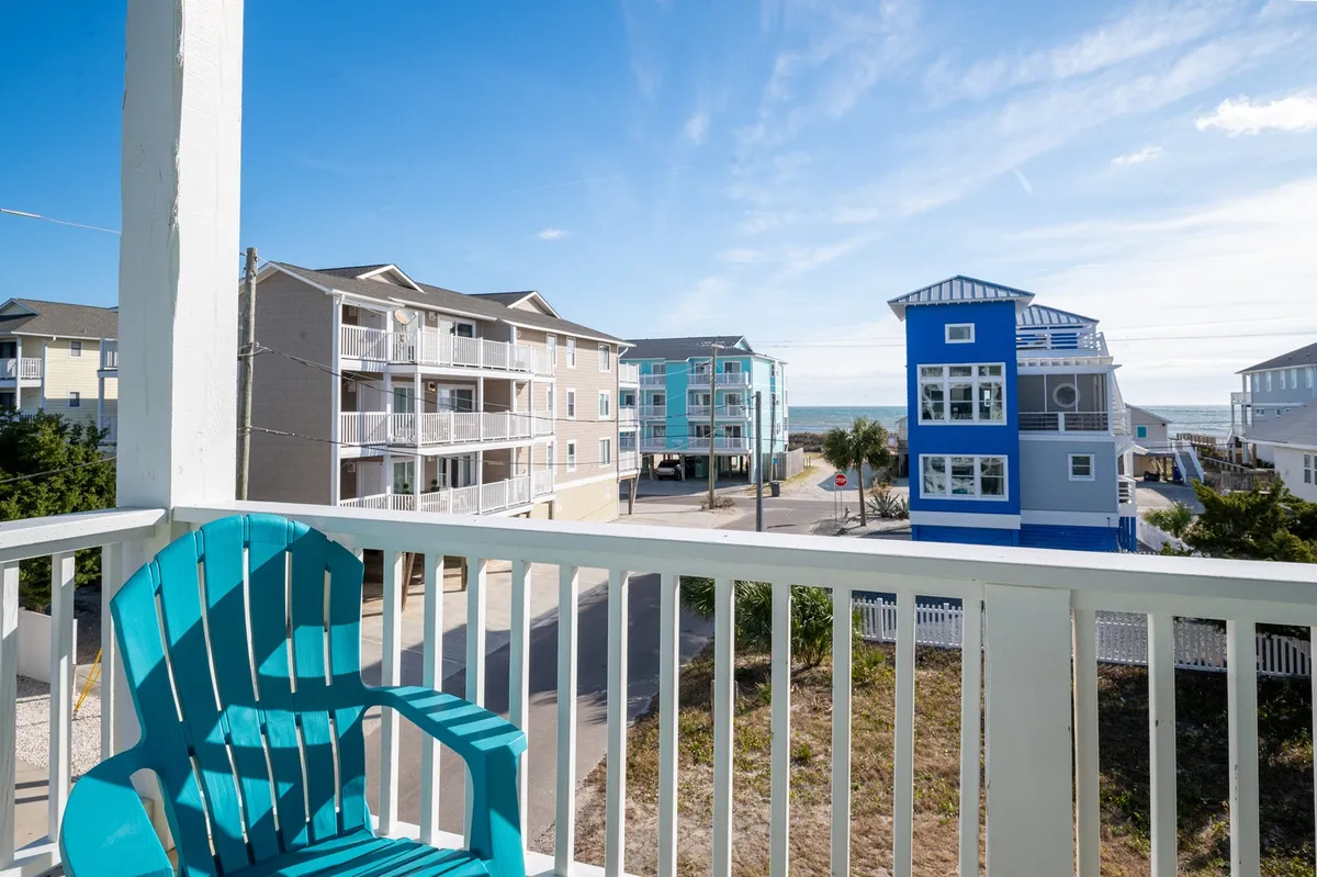 A balcony scene overlooking the beach, featuring a turquoise chair and a blue building in the foreground. The ocean is visible beyond, with open sky and bright surroundings setting a cheerful, inviting mood. The setting suggests prime access to beachfront activities and local events.