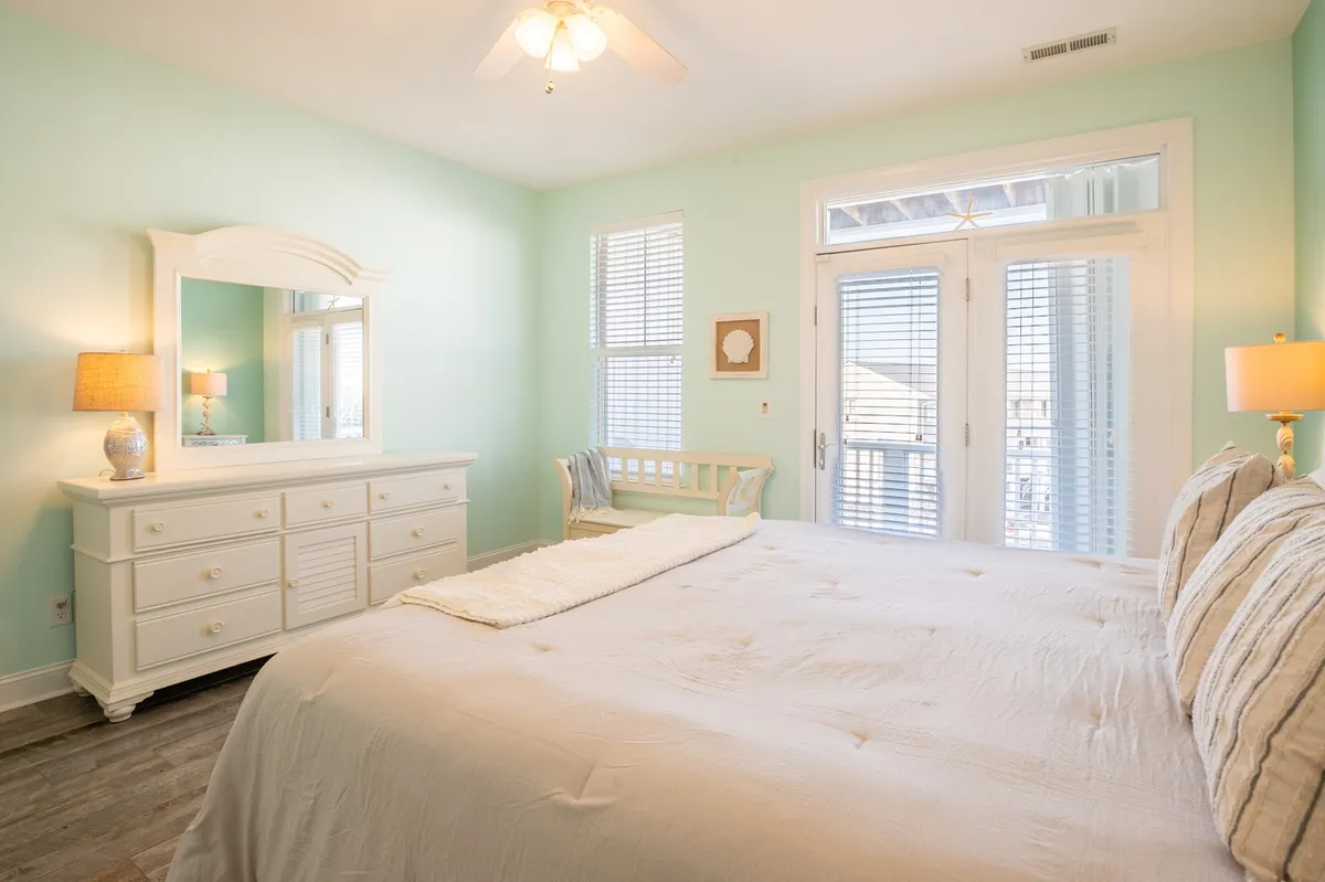 A spacious, sunlit bedroom featuring a large bed with crisp white bedding, a bright dresser with mirror, and double doors that open onto a private balcony. The room is filled with natural light, creating a serene and inviting atmosphere perfect for relaxation and retreat.