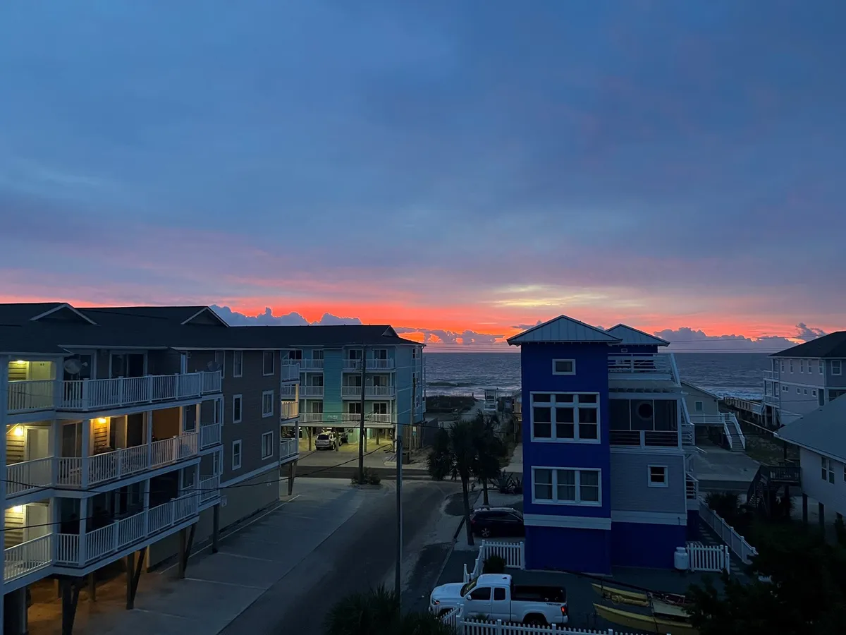 A stunning sunset over the ocean sets the scene, with colorful skies and beachfront buildings stretching along the shoreline. Groups of people can be imagined gathering to watch the sunset, stroll along the beach, or capture photos together.