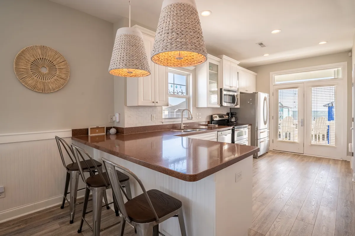 A modern kitchen with a spacious island, hanging light fixtures, stainless appliances, and windows overlooking the outdoors. The layout is open and ideal for both cooking and casual conversations at the breakfast bar.