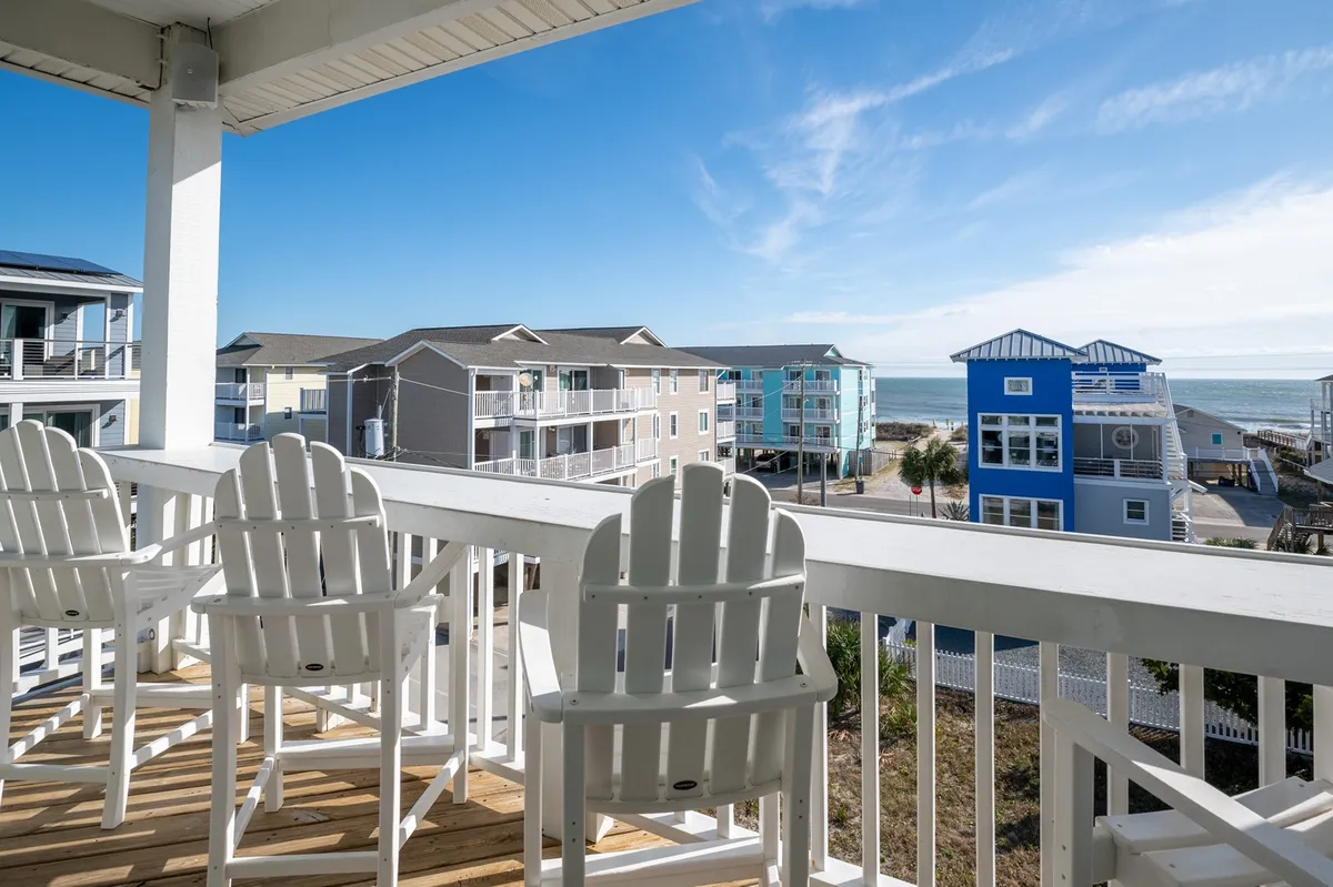 A covered porch with white Adirondack chairs facing a stunning ocean view and colorful buildings. The tranquil setting invites relaxation while soaking in coastal vistas.