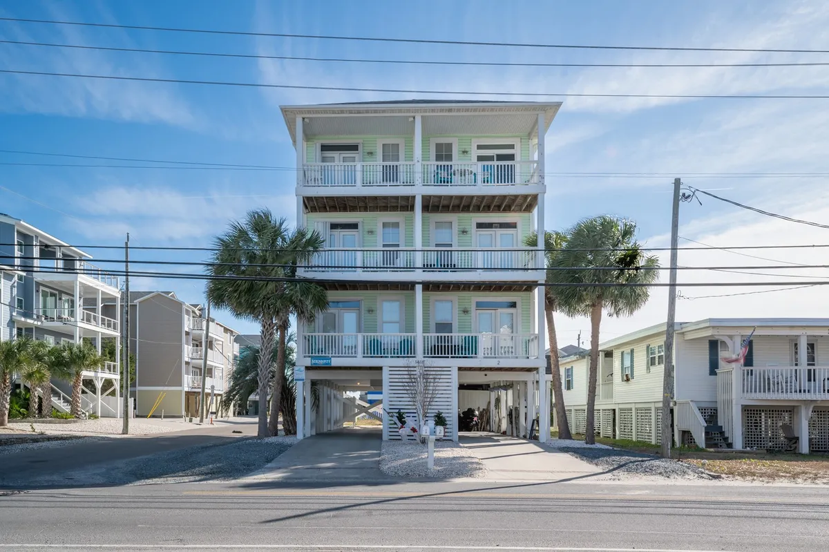 The cheerful exterior of a three-story townhouse framed by lush palm trees, with a driveway and balconies perfect for enjoying the coastal breeze. The home radiates a welcoming, vacation-ready vibe close to the beach.