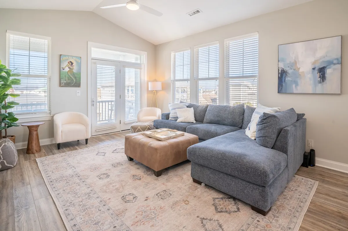 A bright, sunlit living room featuring a large sectional sofa, a central coffee table, tasteful artwork on the wall, and expansive windows that allow natural light to pour in. The arrangement is cozy yet spacious, ideal for lounging or socializing.